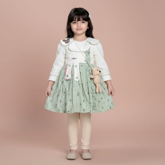 Young girl in a green checkered dress holding a teddy bear, standing between two flower pots against a dark background.